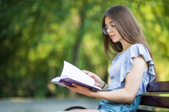 garota de óculos e blusa azul sentada ao ar livre lendo um livro